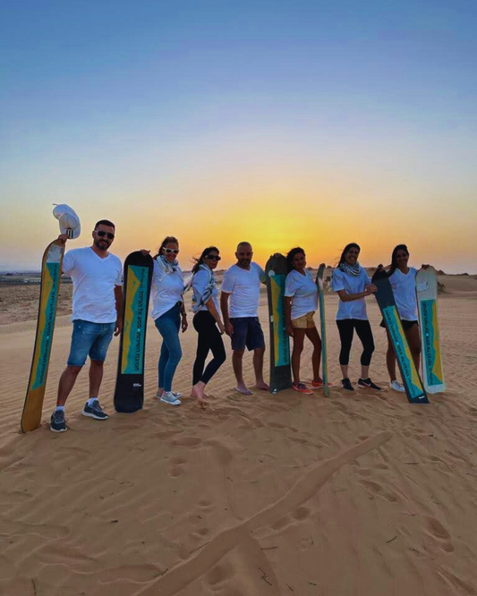 Group of people standing in the desert with sandboards at sunset.